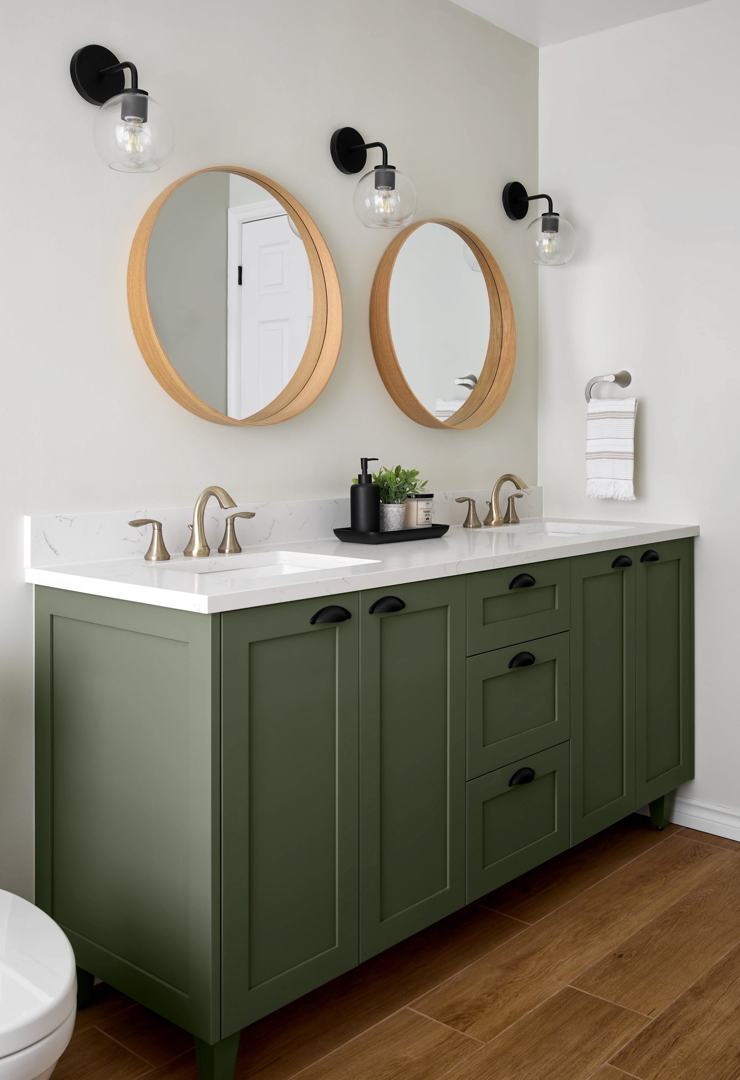 A modern bathroom vanity with a green cabinet, white marble countertop, and two oval wooden-framed mirrors. There are three black wall-mounted light fixtures above the mirrors. The vanity has brass faucets and a black tray with a soap dispenser, hand