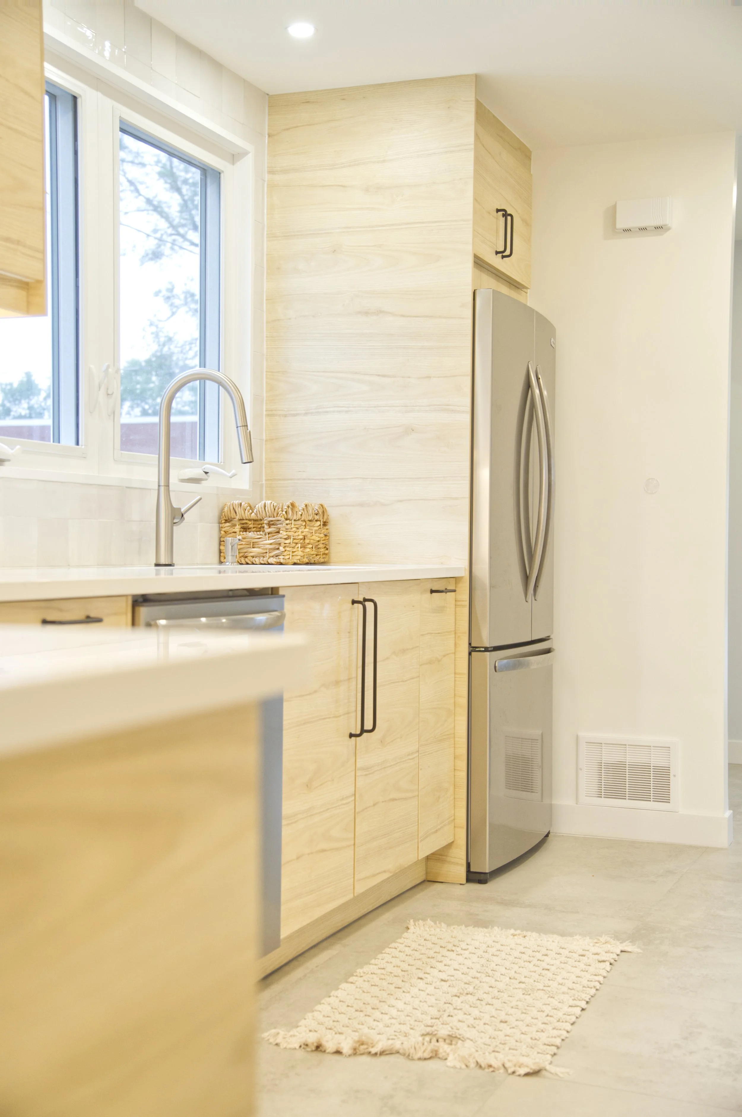 A modern kitchen with light wood cabinets, a stainless steel refrigerator, a white countertop, a woven basket next to a window, and a textured beige rug on the floor.