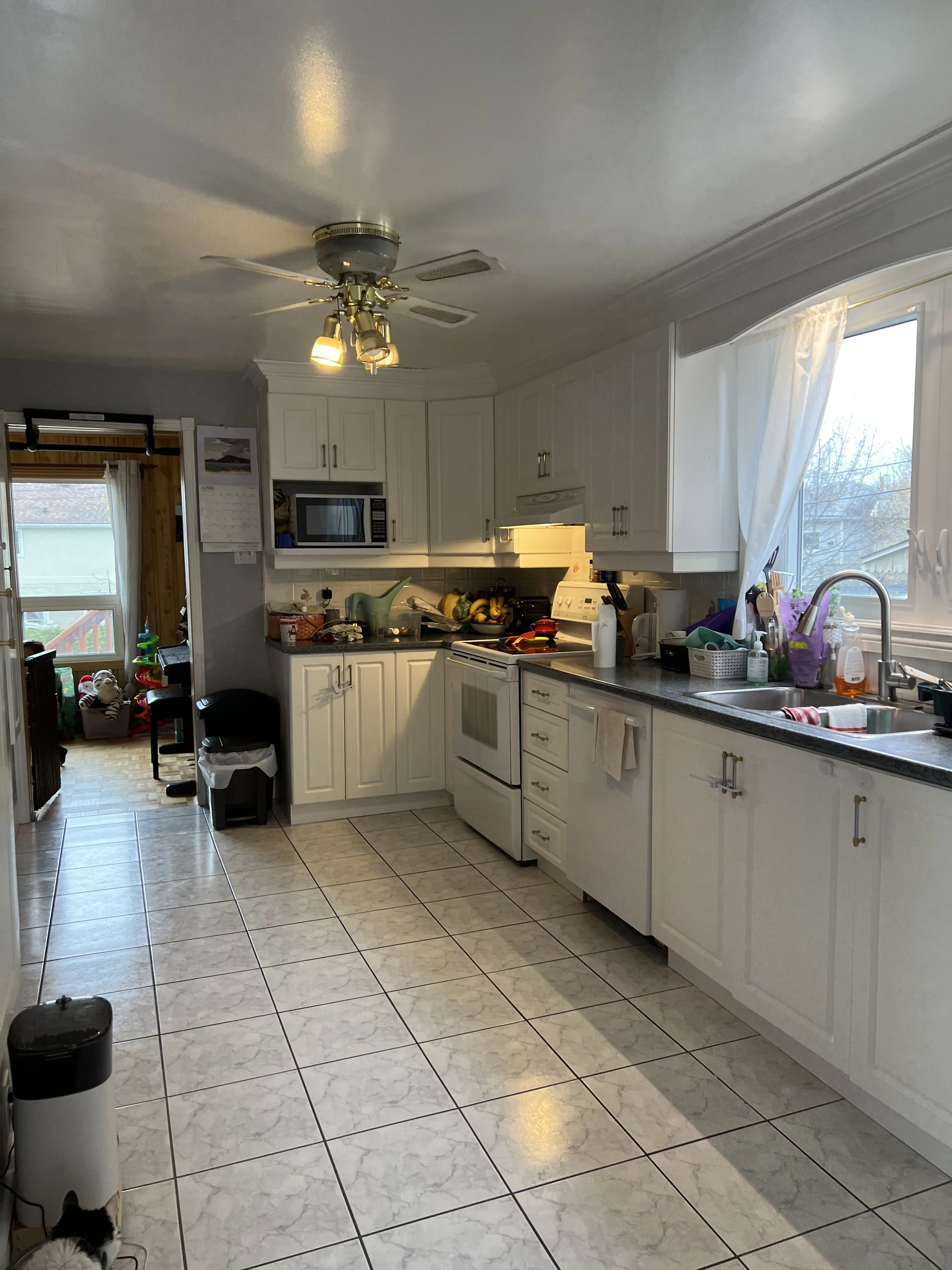 A kitchen with white cabinets, a window with a white curtain, a white stove, and various kitchen items on the counter. There is a ceiling fan with lights and a doorway leading to a sunroom or another room with door and window.