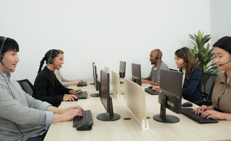Six diverse customer service representatives with headsets working at a shared desk with computer monitors in an office.