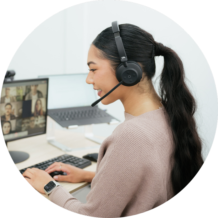 A woman wearing a headset with a microphone, working at a desk with a computer monitor showing a video conference call, in a bright office environment.