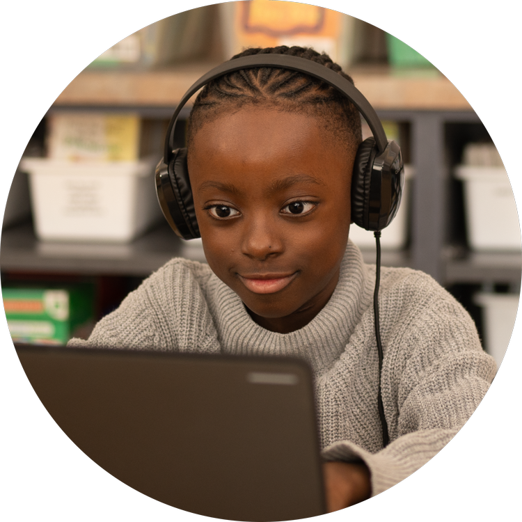 A young girl wearing headphones and a beige sweater, focused on a laptop in a classroom setting
