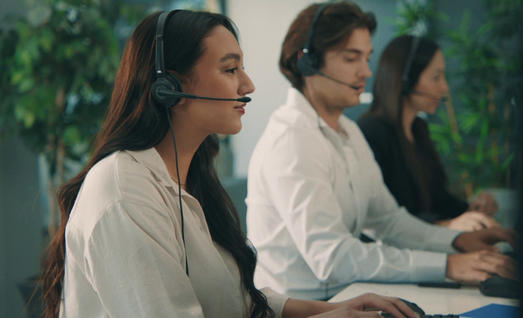 Three customer service representatives wearing headsets, sitting at desks and working on computers, in an office environment with green plants in the background.