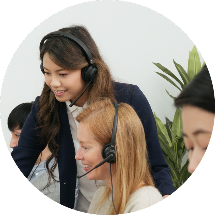 A woman with long brown hair in a navy blazer assisting customer service representatives in headsets at a desk