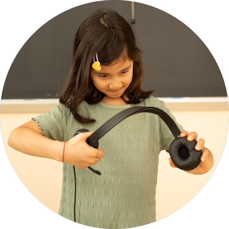 A young girl in a green shirt standing in front of a blackboard holding headphones