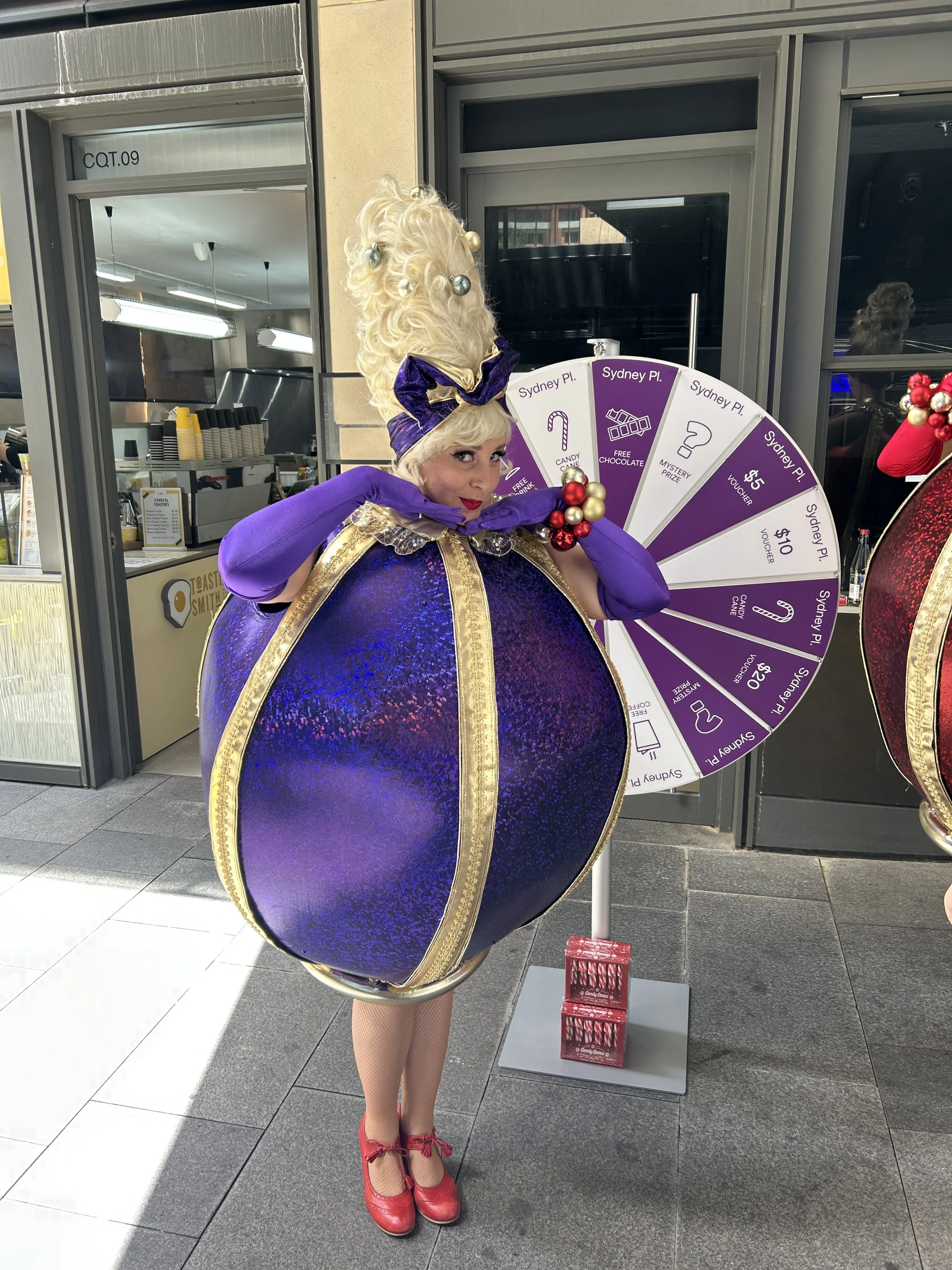 A woman in a ballerina costume designed to look like a large, shiny purple and gold ornament. She is holding a purple glove to her face and standing next to a prize wheel.