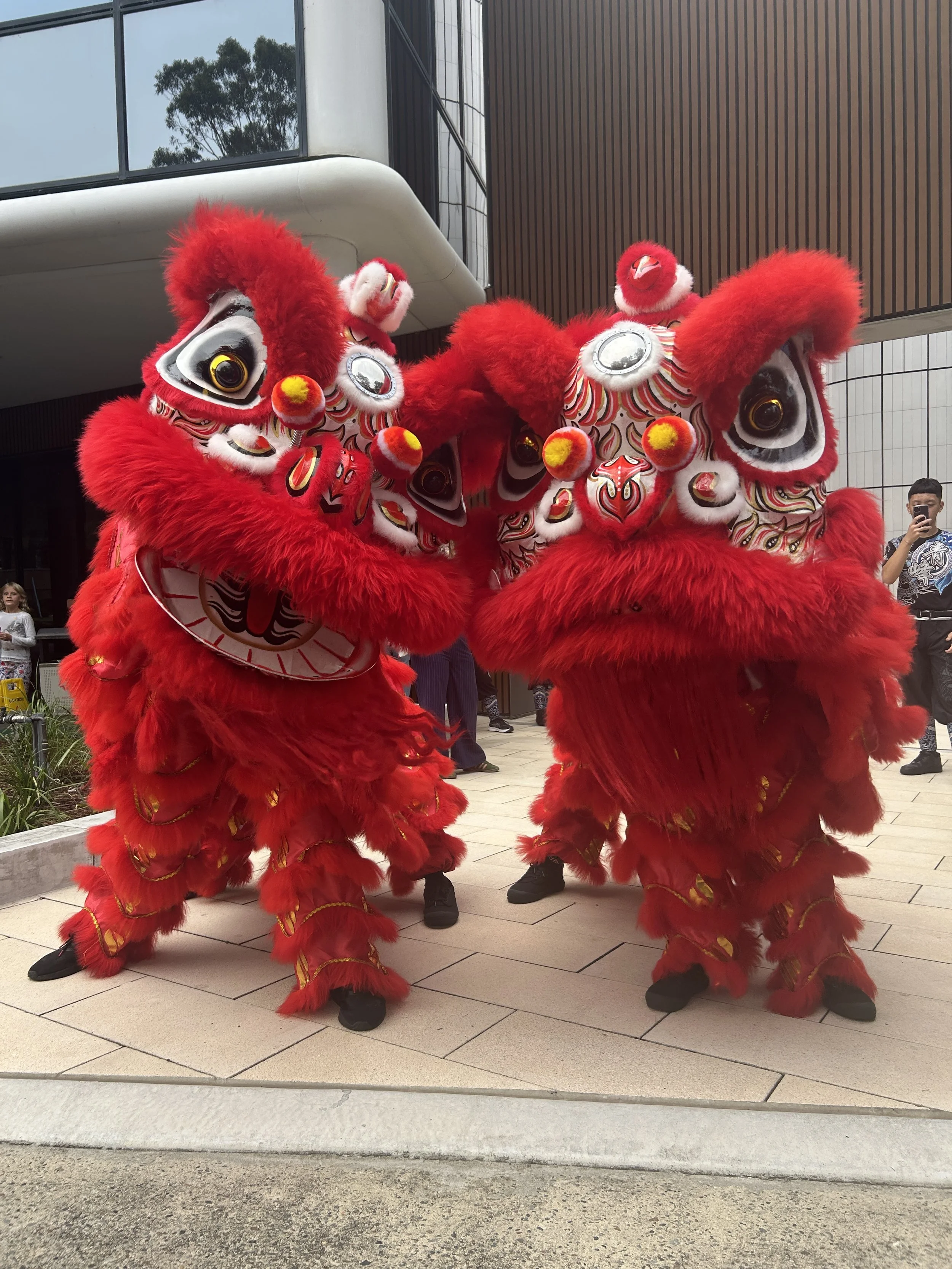 Two performers dressed in red Chinese lion dance costumes with large, expressive eyes and intricate designs, performing outdoors with onlookers and modern buildings in the background.