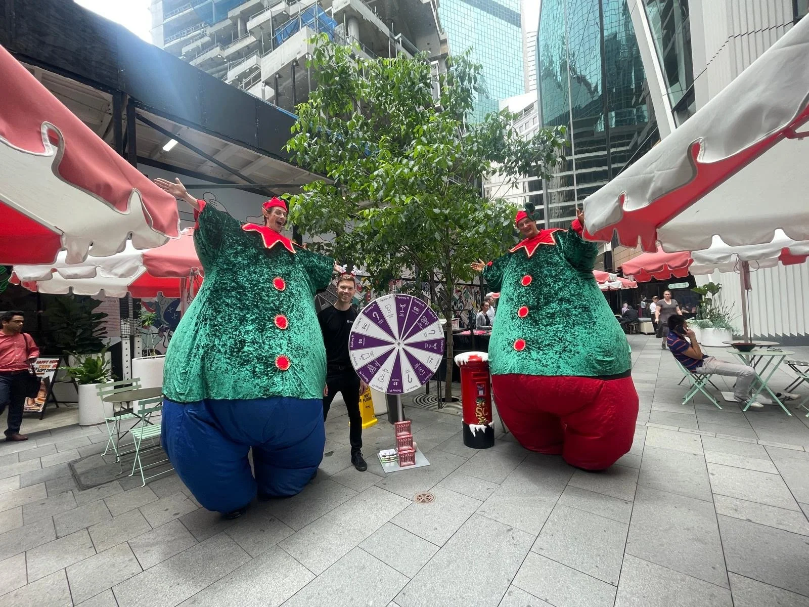 Two people dressed in oversized Christmas elf costumes standing on a sidewalk in a city, next to a man in black, with a wheel of fortune game and a red fire hydrant between them. The scene is outdoors with modern buildings and trees in the background