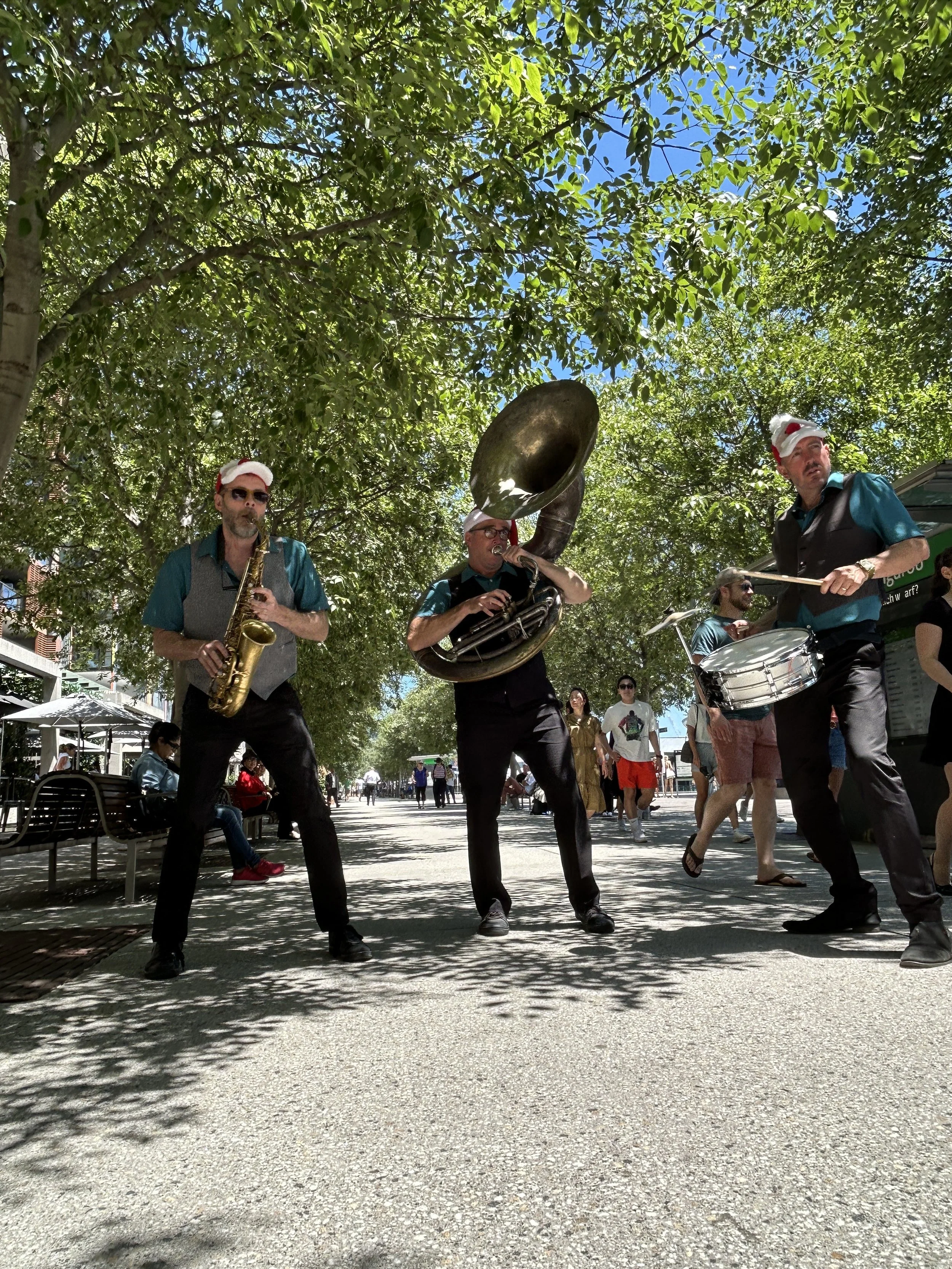 Three street musicians wearing festive Santa hats performing with their instruments on a shaded pedestrian street, surrounded by onlookers and trees.