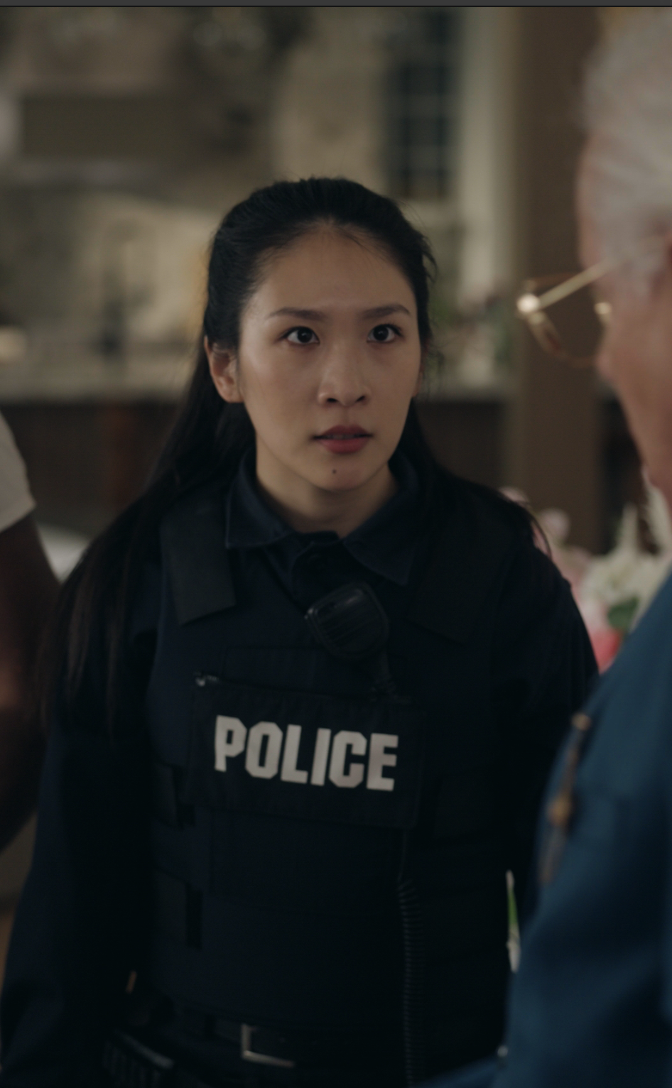 A female police officer wearing a dark uniform and bulletproof vest labeled “POLICE” looks intently at someone off-camera in a home interior setting, appearing serious and focused.