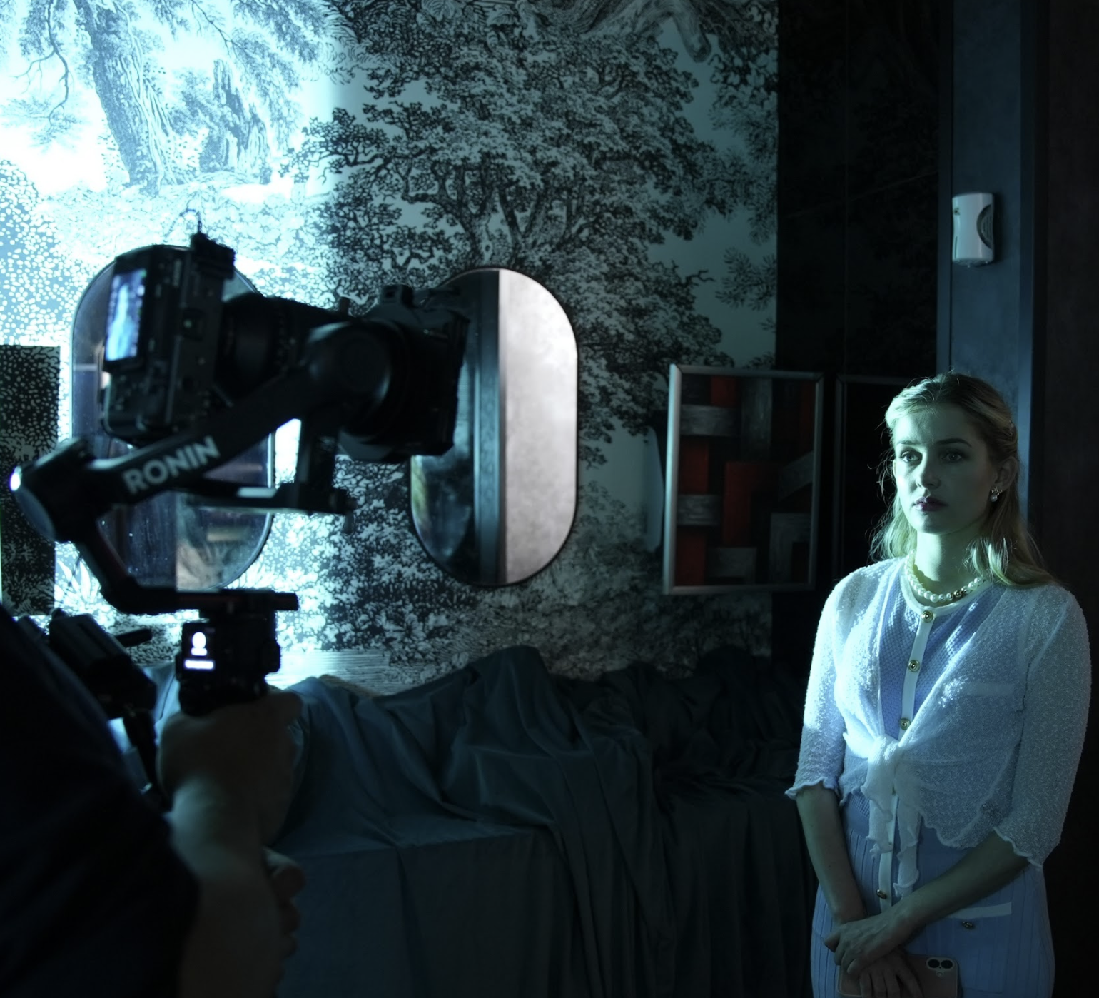 A woman stands against a dramatic, blue-lit backdrop with her hands clasped in front of her while a camera operator films her up close using a mounted camera rig.