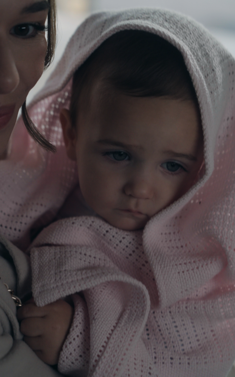 A baby wrapped in a soft pink blanket looks down with a calm, slightly sad expression while being held closely by an adult whose face is partially visible.