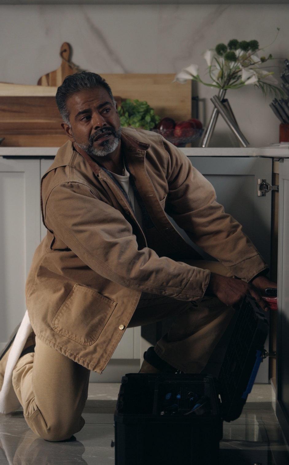 A man wearing a tan jacket and work pants kneels on a kitchen floor, reaching into a toolbox while working on a cabinet. Cutting boards, flowers, and kitchen items are visible on the counter behind him.