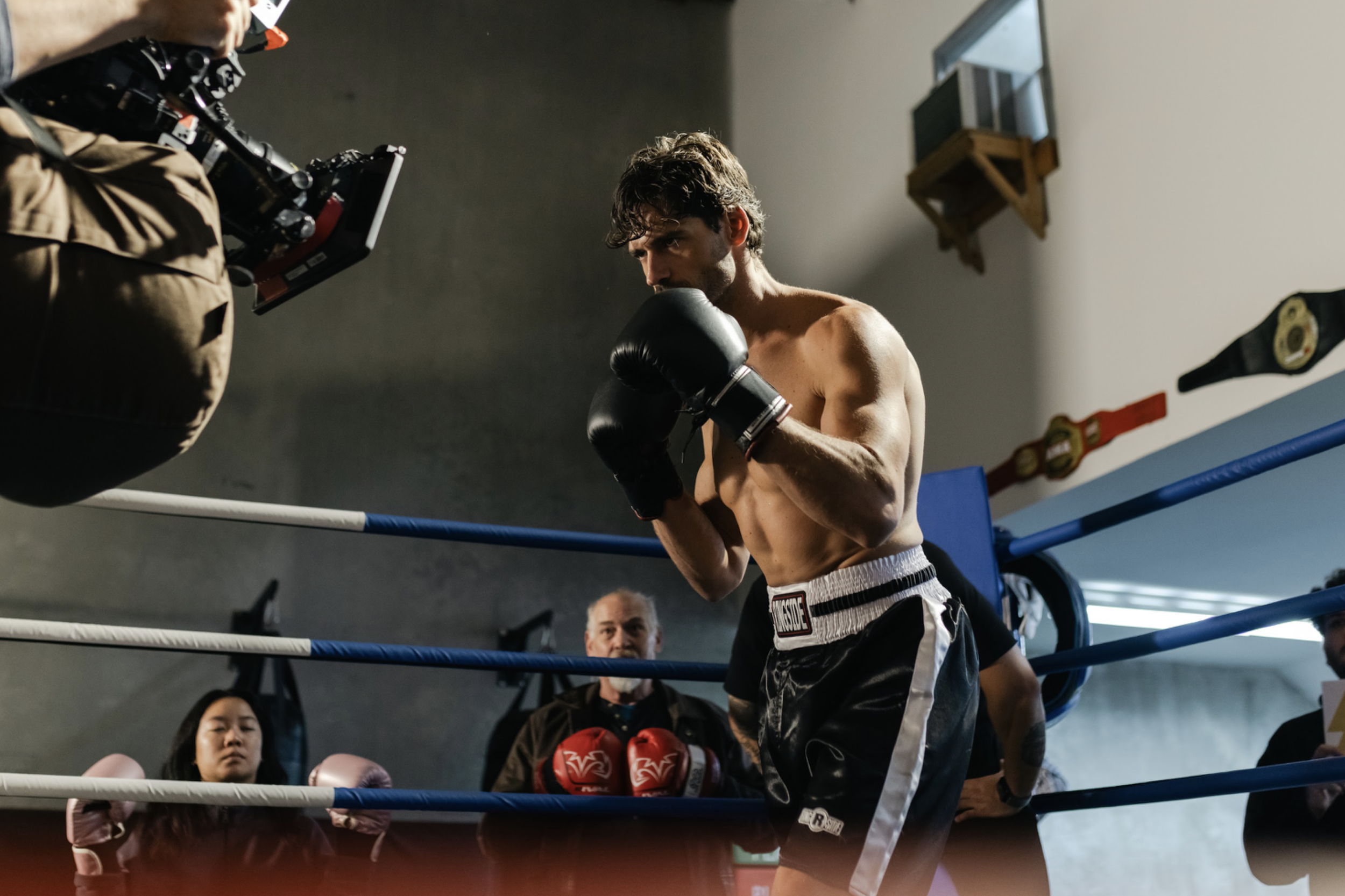 A boxer in black shorts and gloves training in a boxing gym, with a focused expression, inside a boxing ring with spectators and trainers watching.