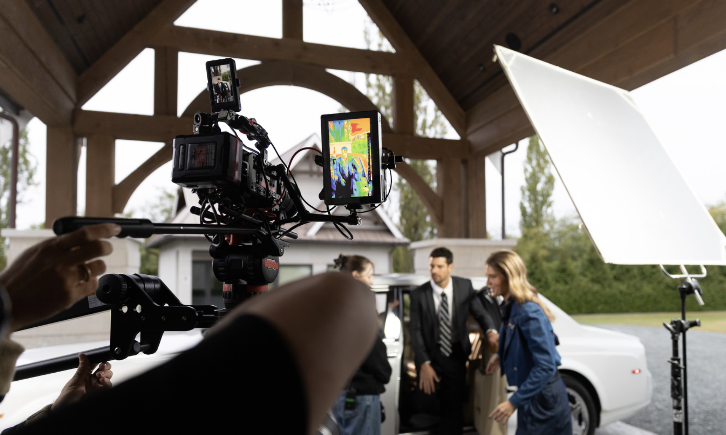 A film crew records a scene beside a white luxury car under a wooden structure. A professional camera rig with monitors is in the foreground, while actors in business and casual attire stand near the car, with lighting equipment set up beside them.