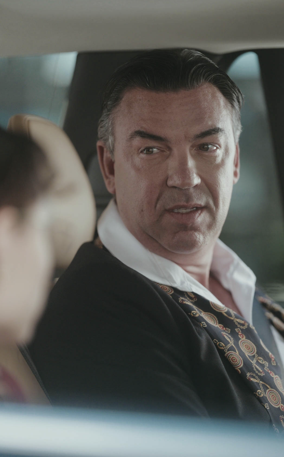 A man in a suit with a patterned collar sits in a car and looks toward someone in the passenger seat, appearing serious and engaged in conversation.