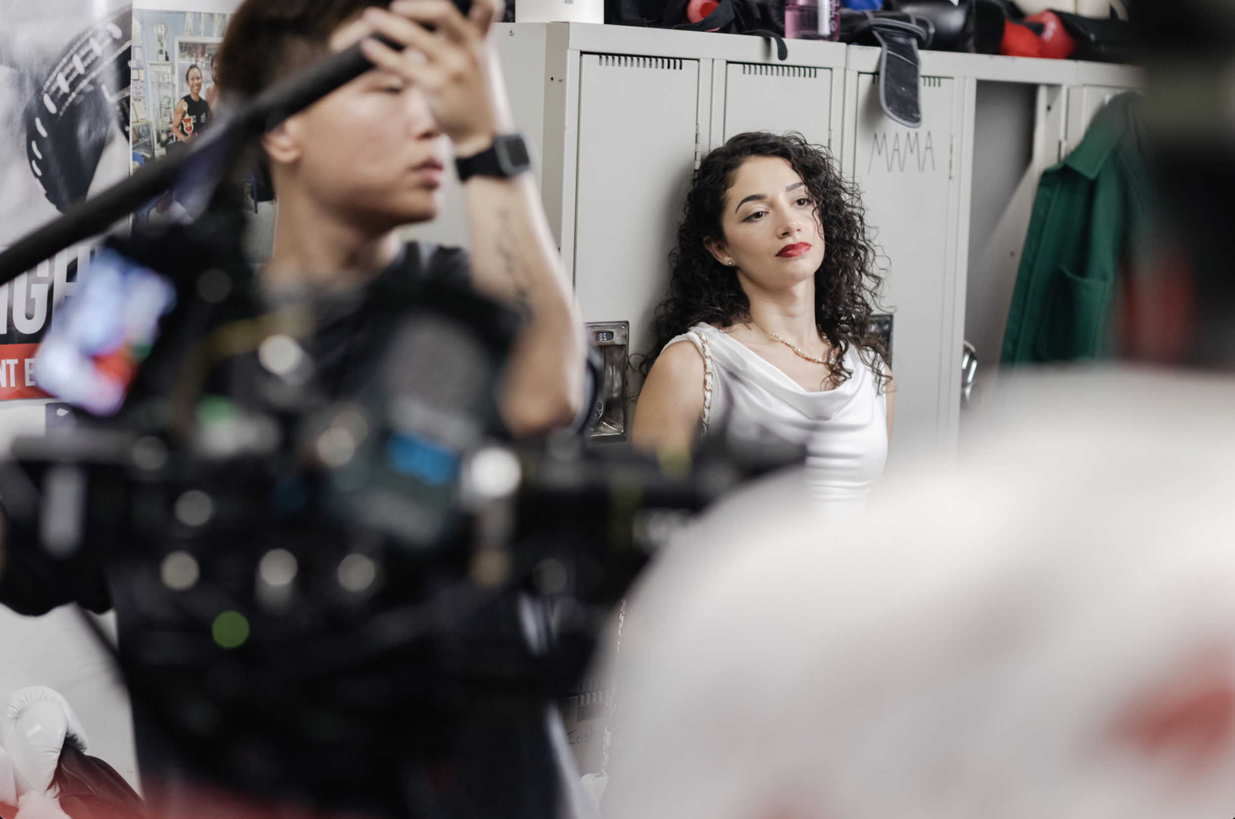 A woman with curly hair in a white outfit stands against a row of lockers on a film set, looking off to the side with a serious expression. In the foreground, a camera operator and equipment partially block the view, showing active filming in progres