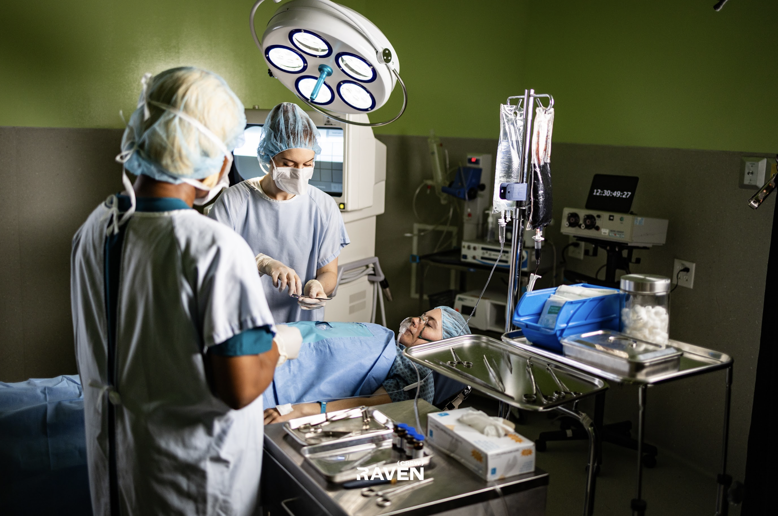Operating room with three medical professionals around a patient lying on the operating table, connected to monitoring equipment. Medical tools and supplies are on nearby tables, with surgical lights overhead and a green wall background.