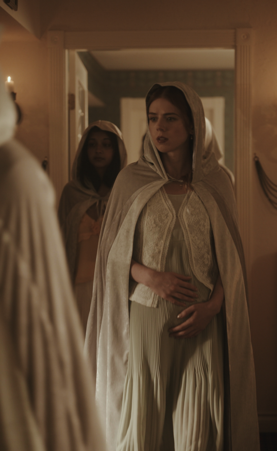 Two women wearing light-colored hooded cloaks stand in a softly lit hallway, with the woman in front holding her hands near her stomach and looking to the side with a concerned expression. Another woman is partially visible behind her.