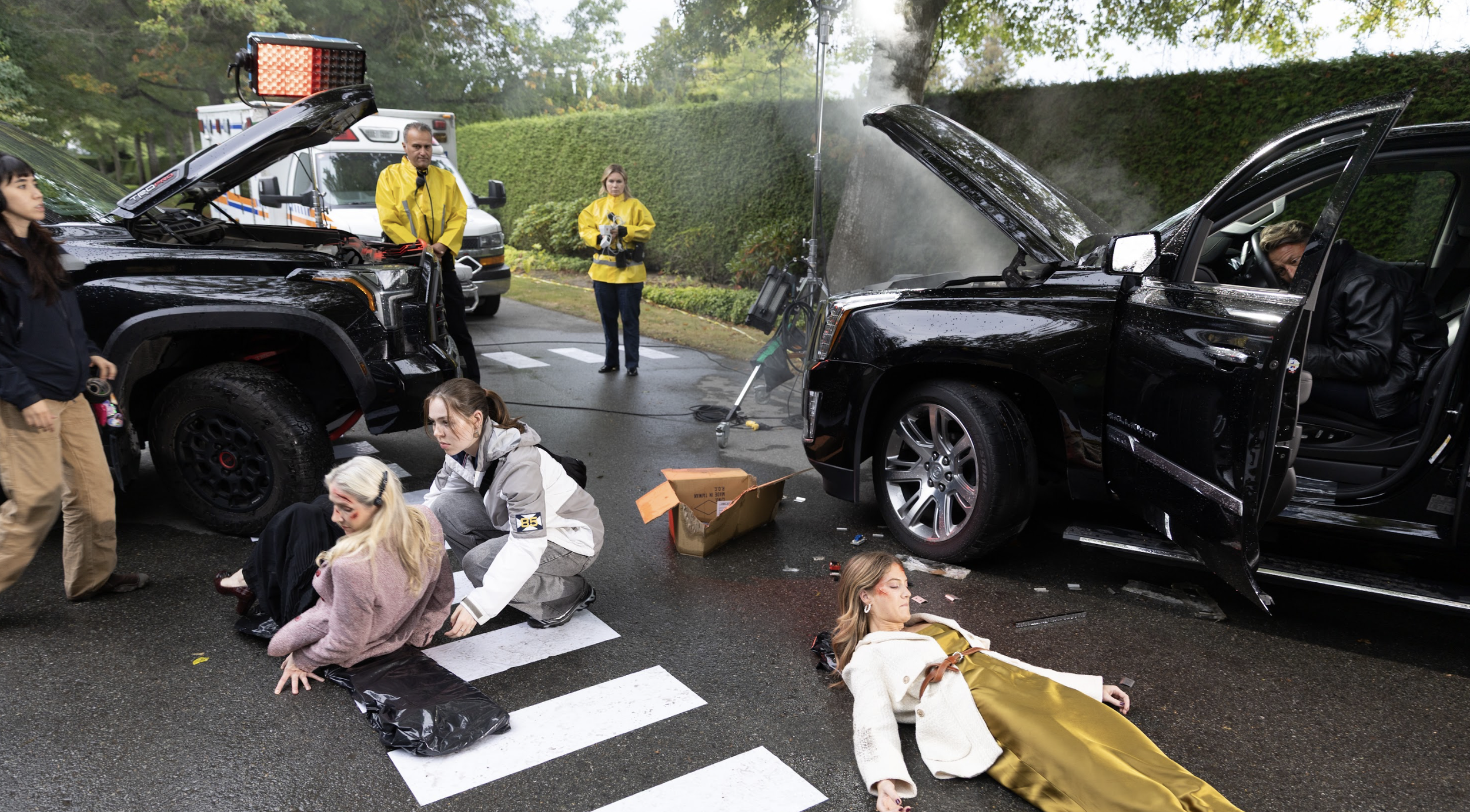 A staged car accident scene on a street shows two black vehicles with open smoking hoods. Actors portraying injured people sit or lie on the ground near a crosswalk, while crew members and responders in yellow jackets stand nearby. Equipment and an a