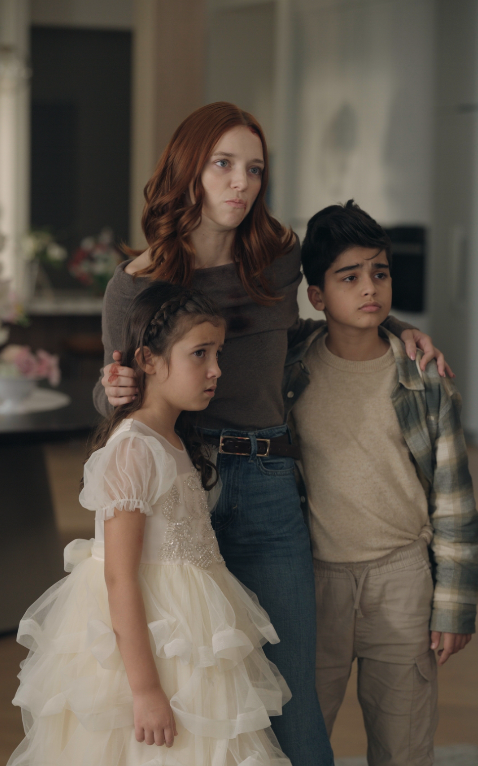 A woman with long red hair stands in a kitchen with her arms protectively around two children, a girl in a white dress and a boy in casual clothes. All three look concerned and are focused on something off-camera.