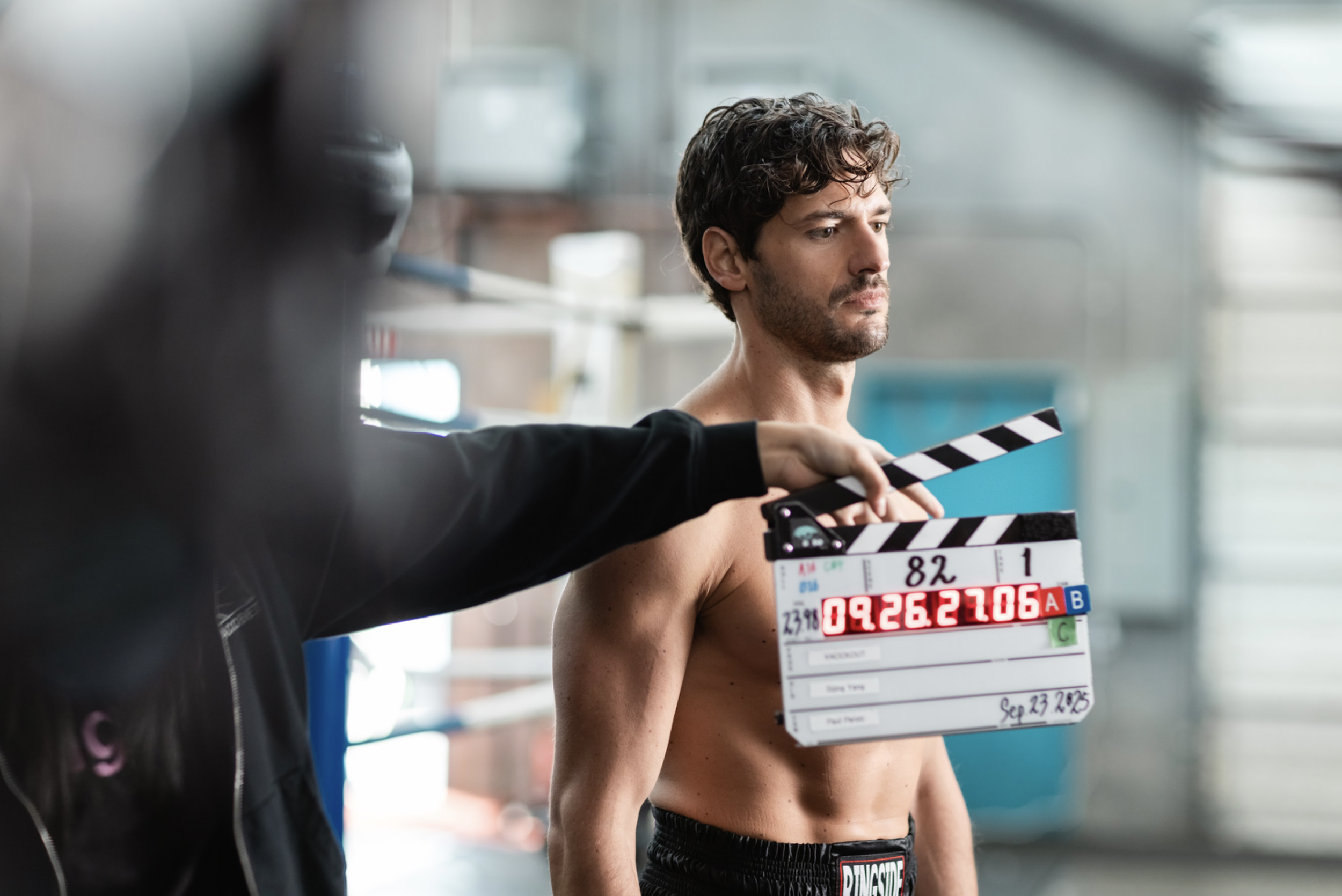 A shirtless male boxer standing in a gym during a film shoot, with a film clapperboard held in front of him.