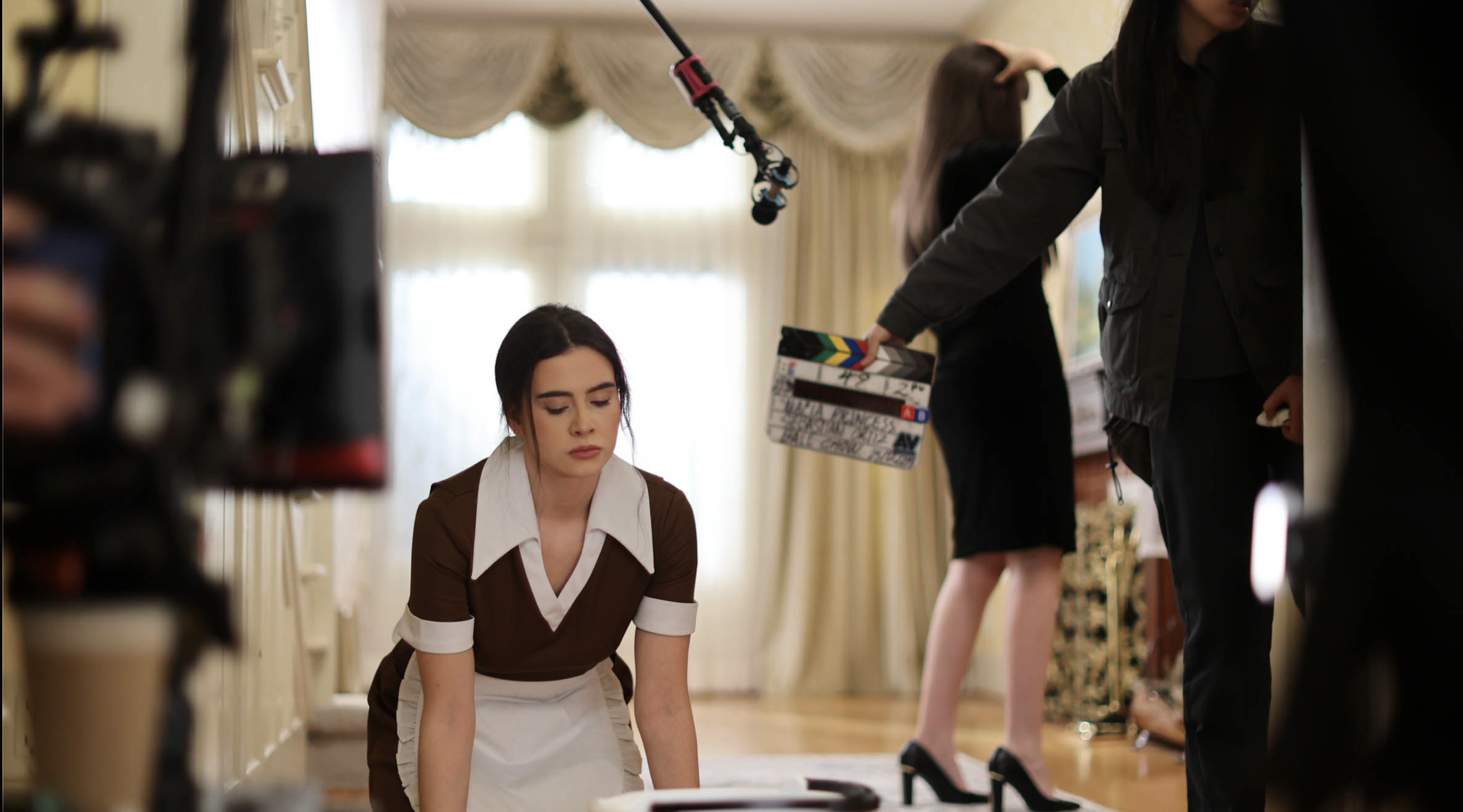 A young woman dressed as a maid kneels on the floor during a film shoot, with a boom microphone overhead and a crew member holding a clapperboard nearby. Camera equipment and another actor in high heels are visible in the background of the elegant ro