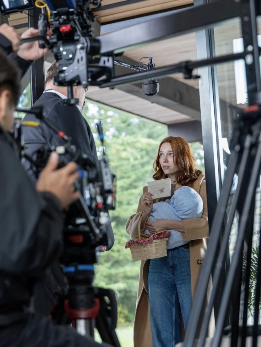 A woman with red hair stands on a film set holding a bundled baby and a small basket, showing an envelope to another actor. Camera equipment and crew are visible around them during the outdoor shoot.