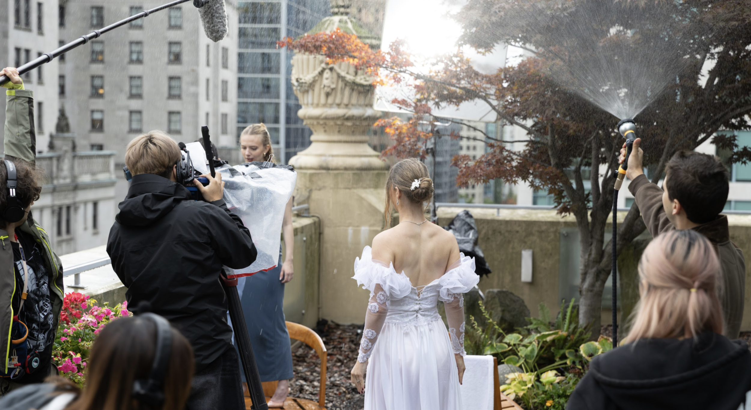 A film crew is shooting a scene on a rooftop garden with city buildings in the background. A woman in an elegant white dress stands in the center facing another actress, while crew members operate a camera, boom microphone, and spray water to simulat