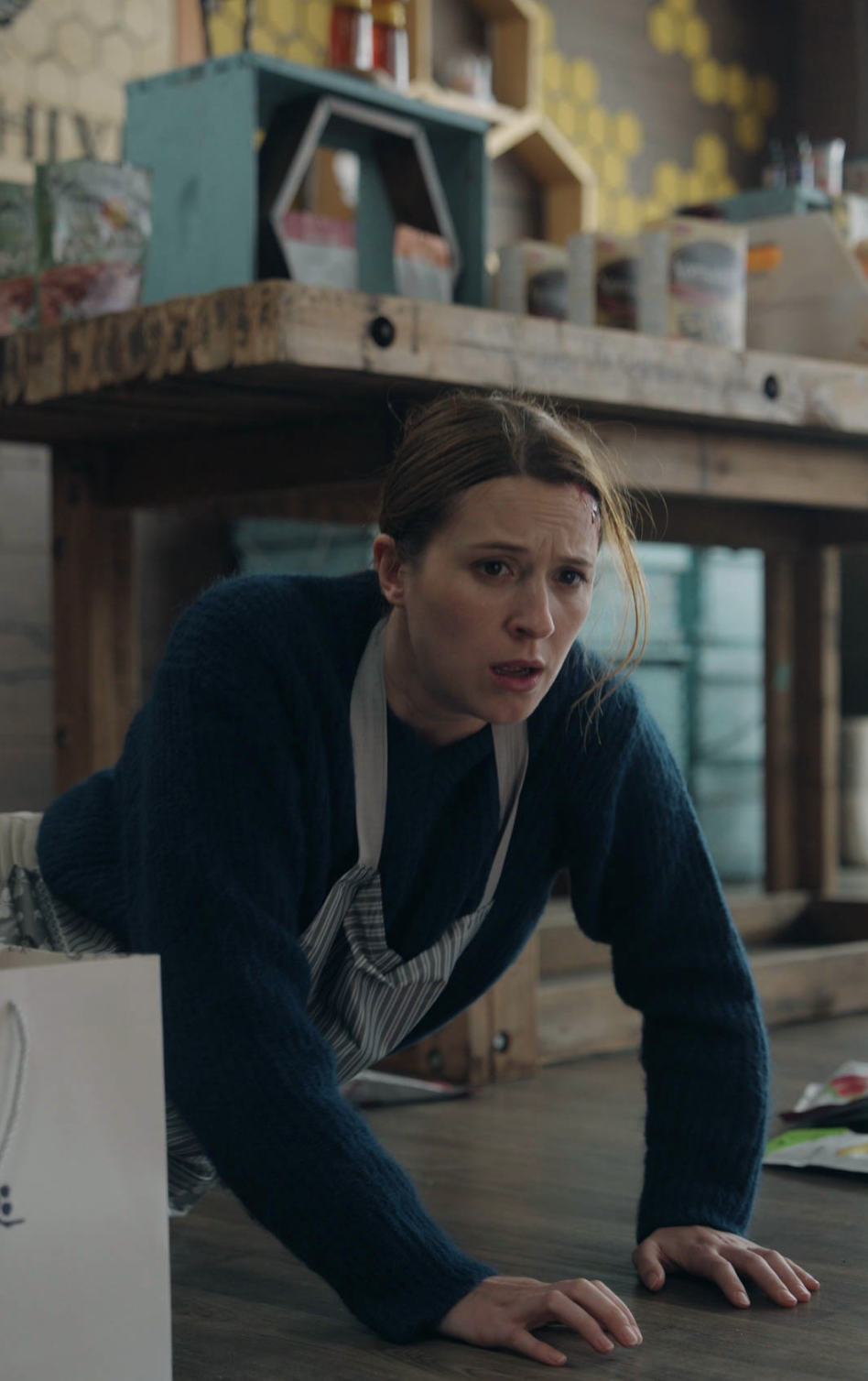A woman wearing a blue sweater and apron is on her hands and knees on the floor of what appears to be a shop or café, looking up with a startled or worried expression. Shelves and supplies are visible behind her.