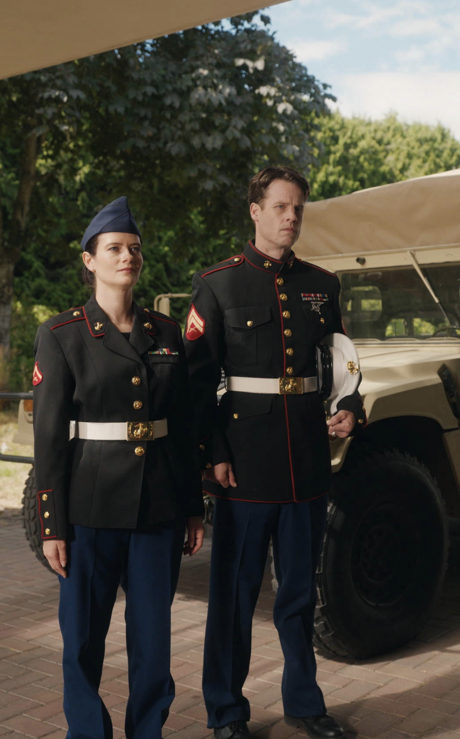 A woman and a man dressed in formal military uniforms stand side by side outdoors near a tan military vehicle, looking ahead with serious, composed expressions.