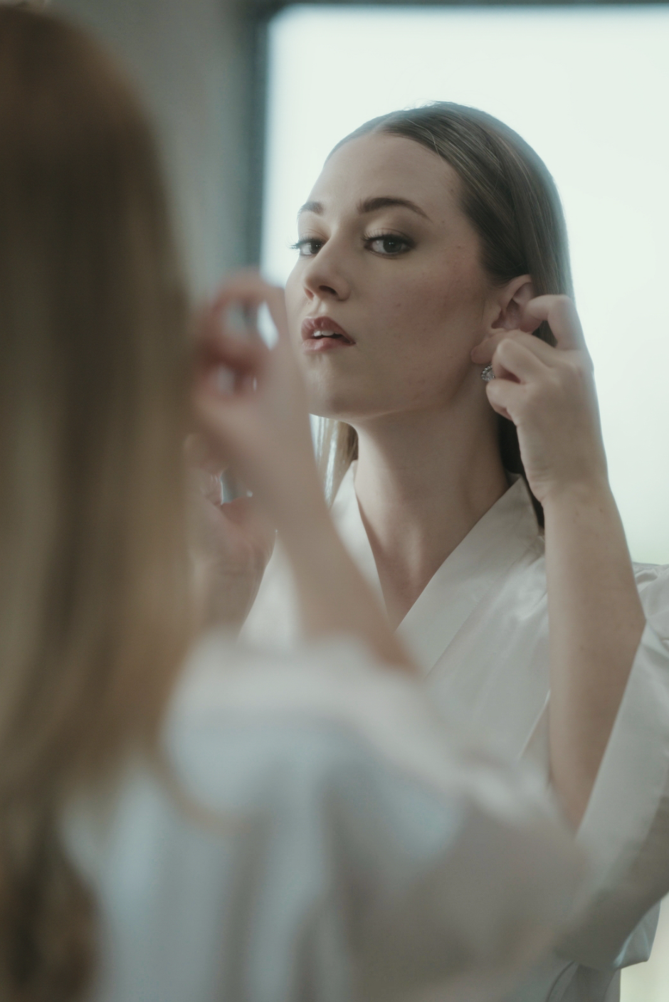 A woman wearing a white robe stands in front of a mirror, adjusting an earring while looking at her reflection with a focused expression.