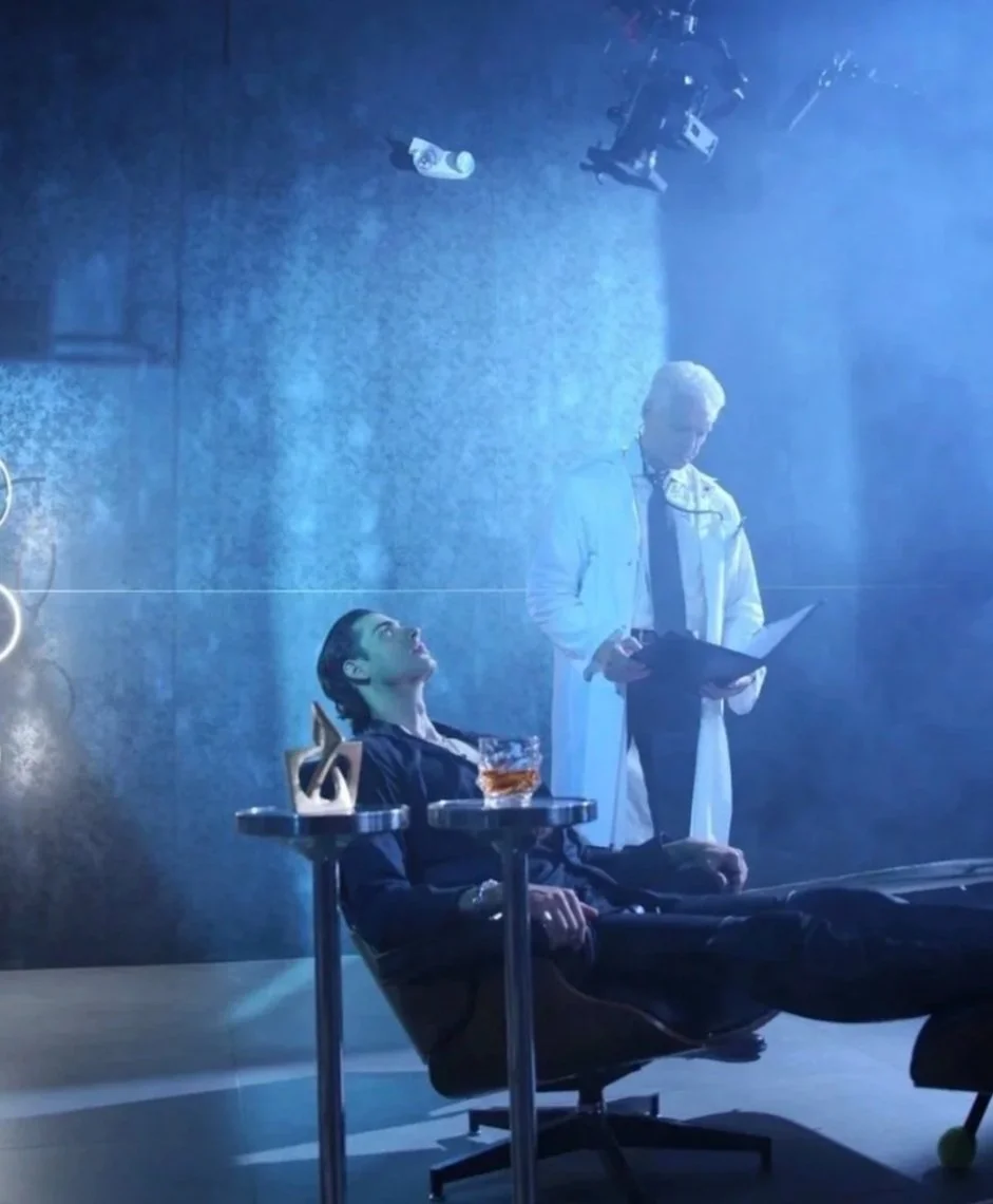 A man sitting in a reclining chair with drink on the table next to him and a male doctor in white gown holding a medical file with a camera rig above them.