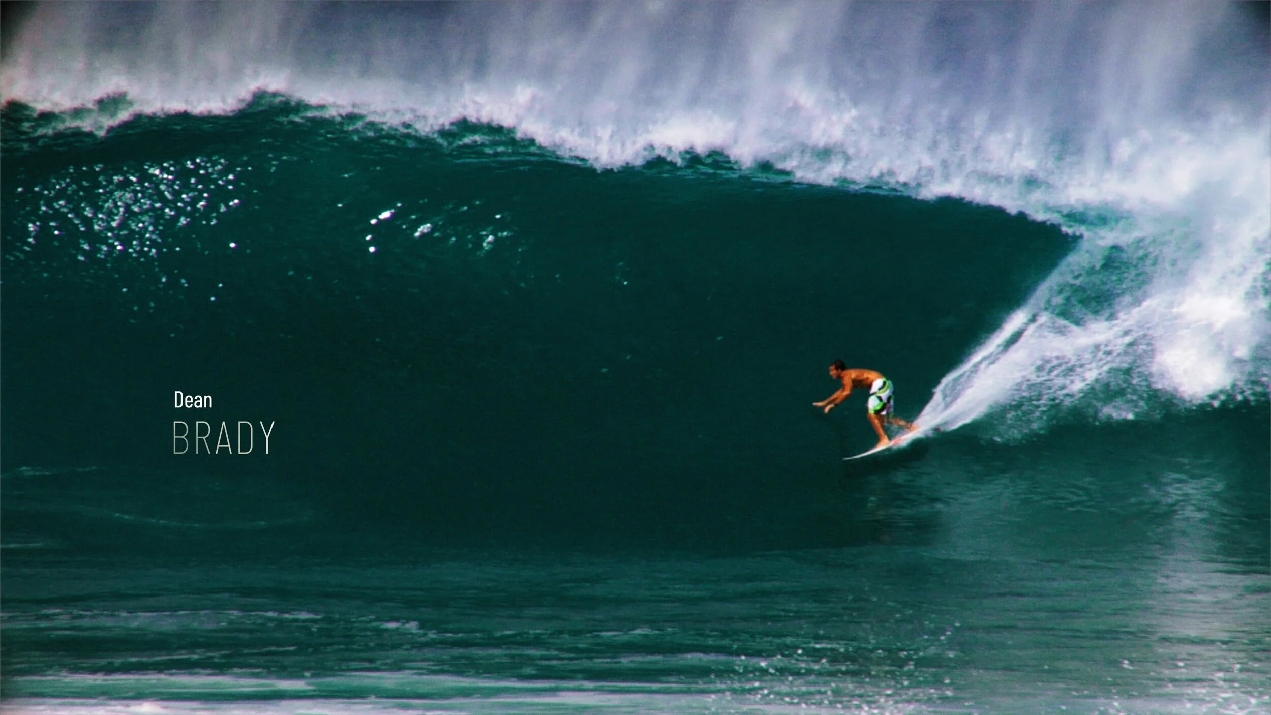 A person surfing on a large ocean wave, wearing swim trunks with green, white, and black patterns, on a white surfboard, with water splashing behind them.