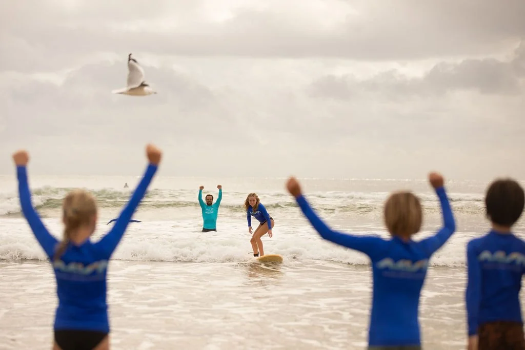 Children and adults at the beach, with children in the foreground raising their arms, and two people in water, one on a surfboard, celebrating or cheering, under cloudy sky.