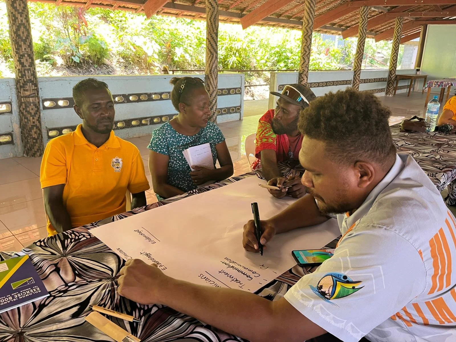 Four GEDSI workshop participants sit around a table under an open-air shelter. One person writes on a large sheet of paper while the others discuss and share ideas.