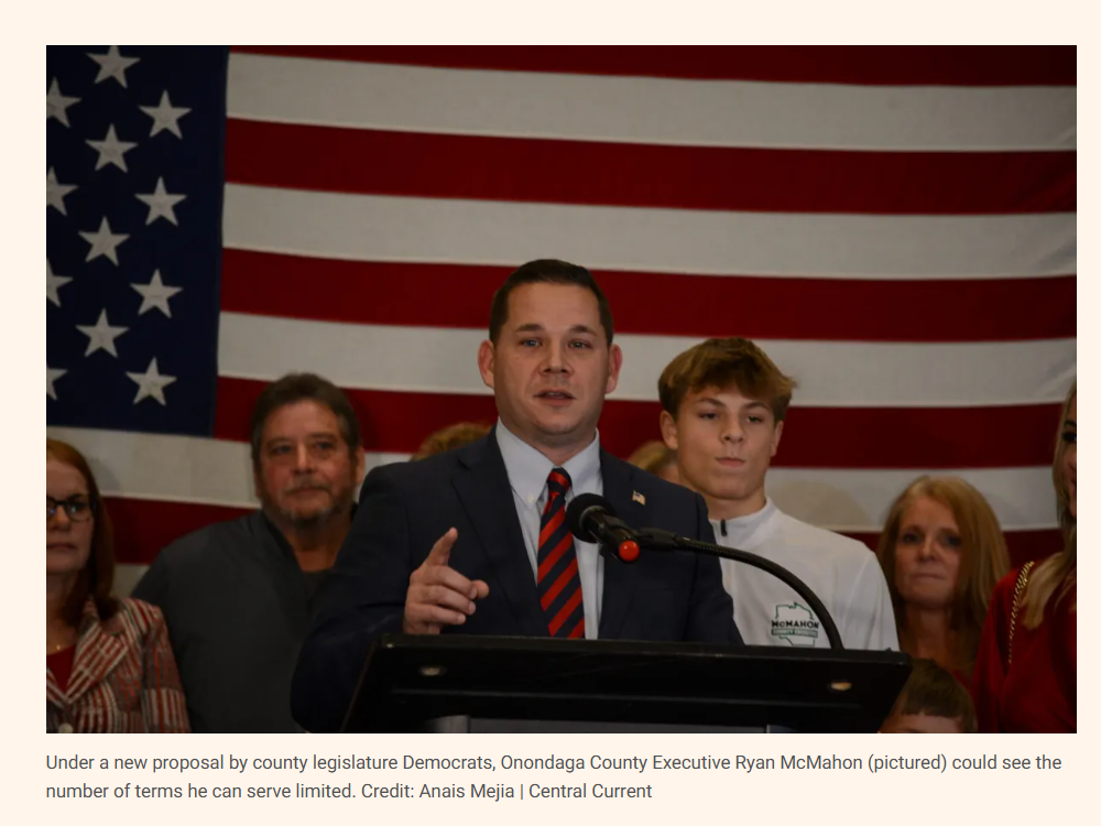 A man in a suit and tie speaks at a podium with a group of people behind him, in front of an American flag