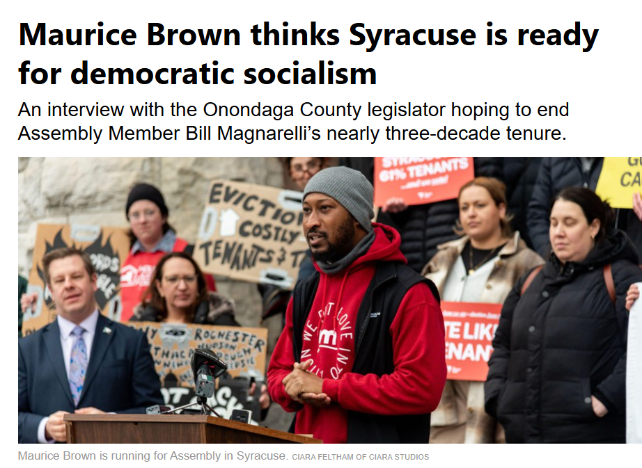 Maurice Brown speaking at a rally in front of a crowd holding protest signs, with a man in a suit standing at a podium.