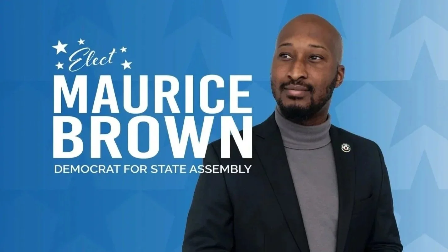 A man with a beard and bald head, wearing a black suit jacket and gray turtleneck, standing against a blue background with white text that says, 'Elect Maurice Brown, Democrat for State Assembly'.