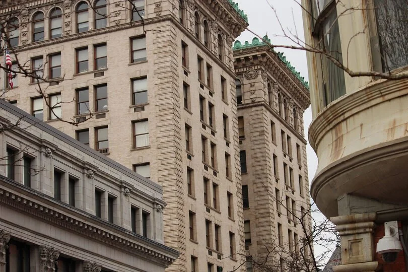 Close-up of historic beige stone buildings with ornate architectural details on a city street, with leafless tree branches in the foreground.