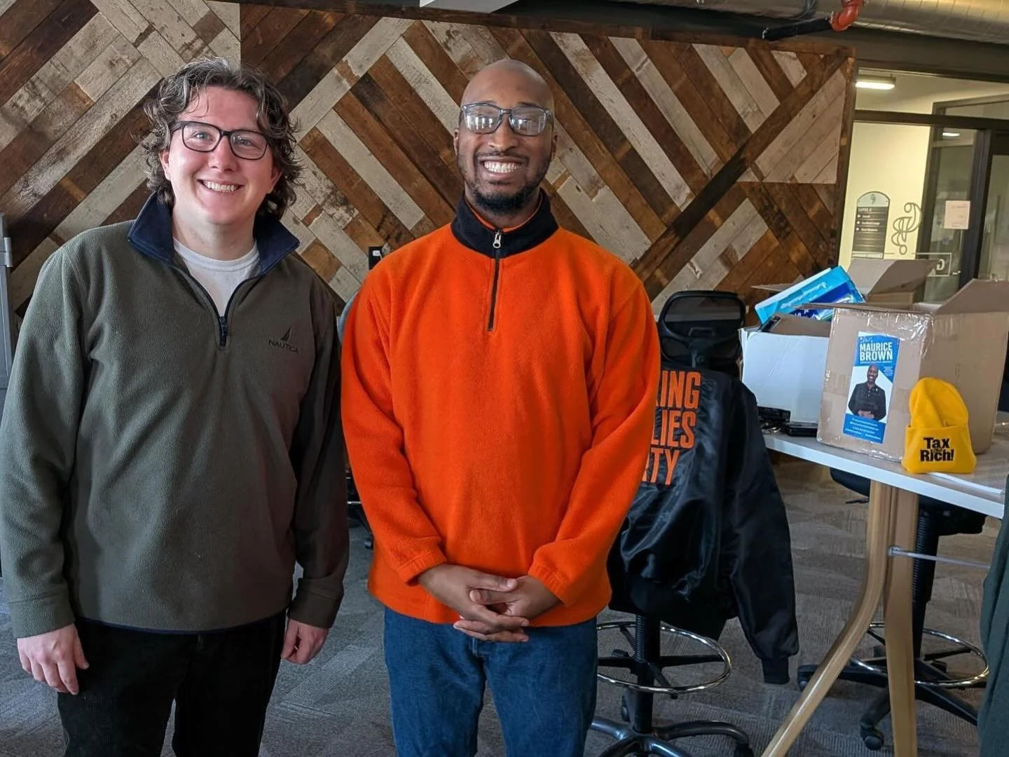 Two men standing indoors, smiling, with a wooden wall background and a table with boxes and promotional materials.
