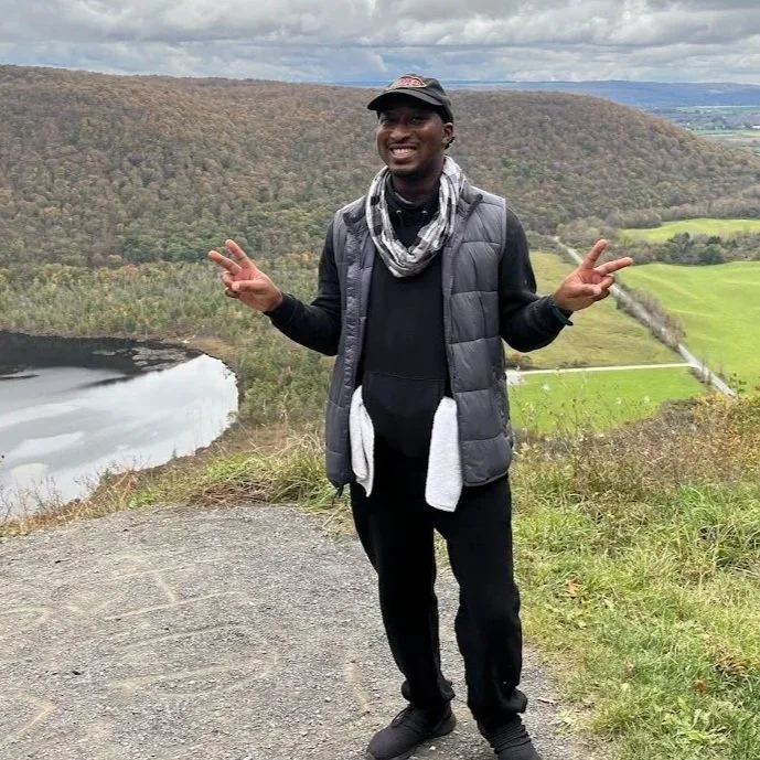 A person smiling and making peace signs with both hands, standing outdoors on a hilltop with a scenic view of a lake, trees, and fields in the background.
