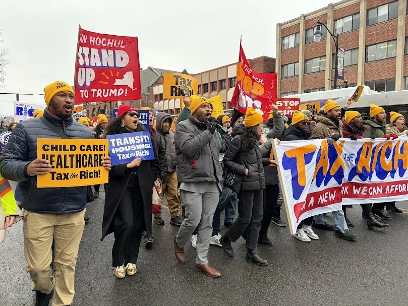 Group of protesters holding signs and banners advocating for taxing rich, healthcare, child care, and transit in a city street.