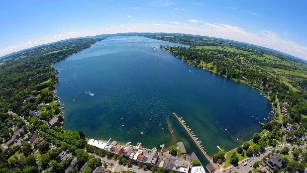 Aerial view of a large river with boats, surrounded by green trees and residential areas under a partly cloudy sky.