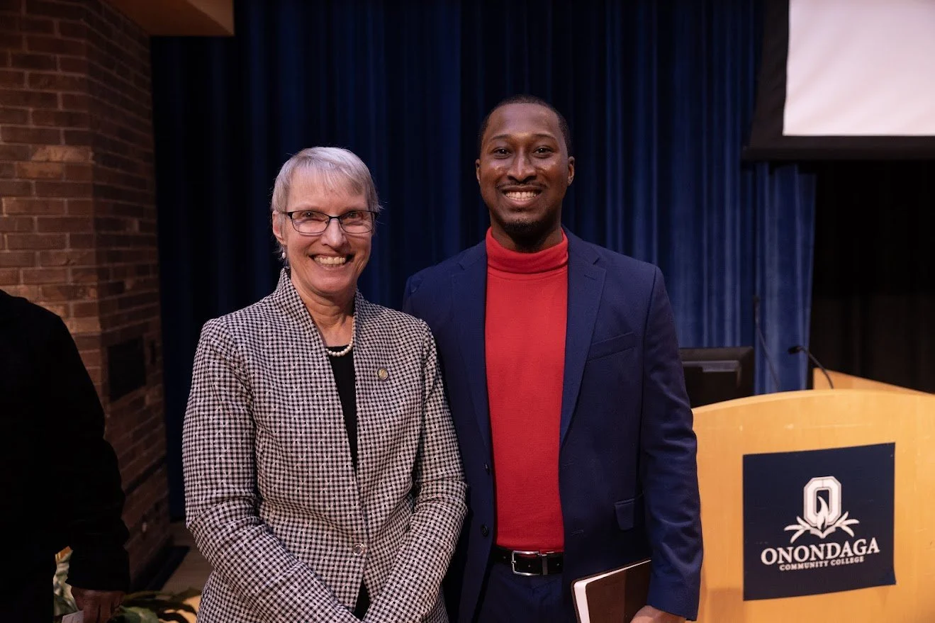 Two people standing indoors, smiling at the camera, with a blue curtain and a podium with the Onondaga Community College logo in the background.