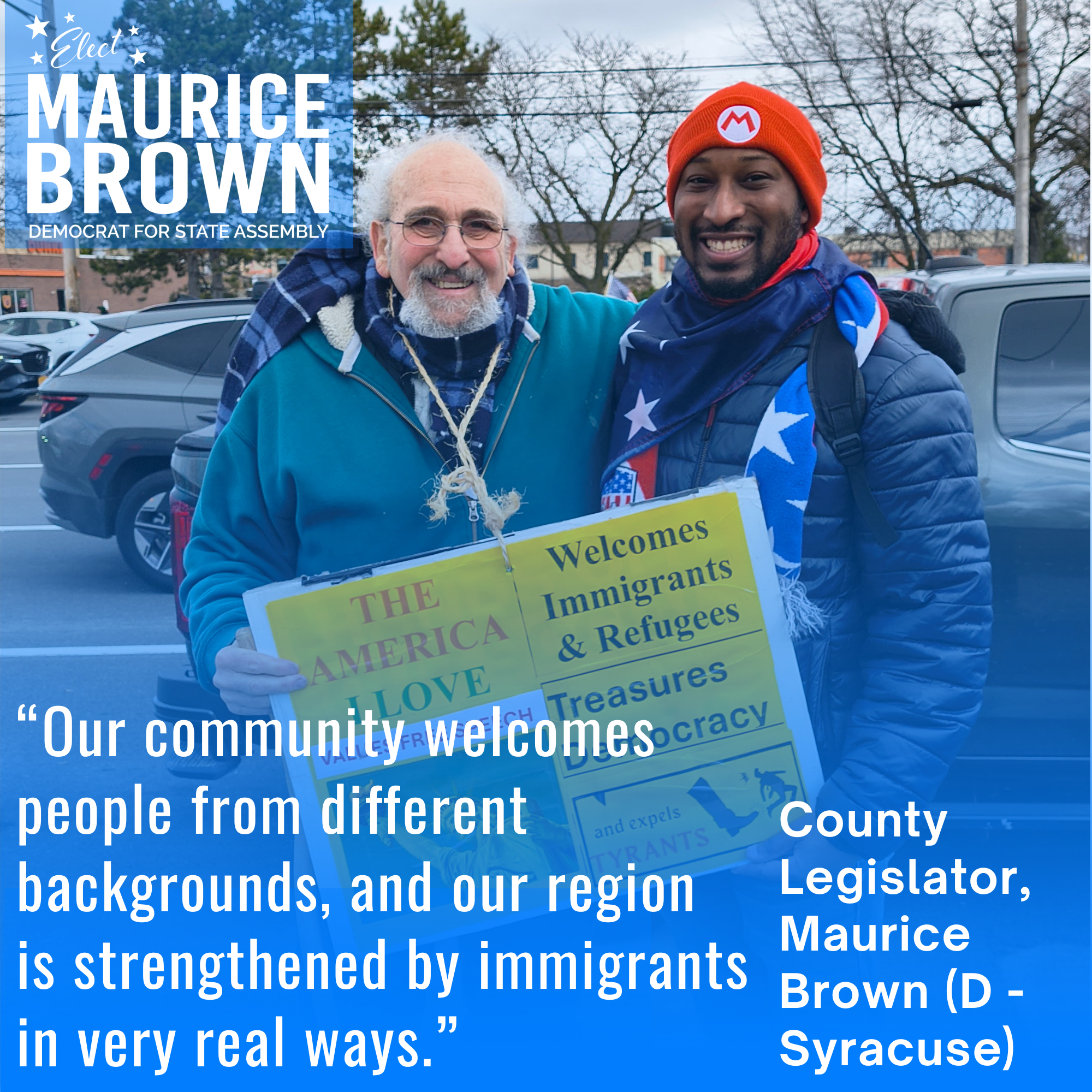 Two men standing together outdoors, one holding a sign that says 'Welcomes Immigrants & Refugees' and other supportive messages, in a parking lot with cars and trees in the background, during daylight.