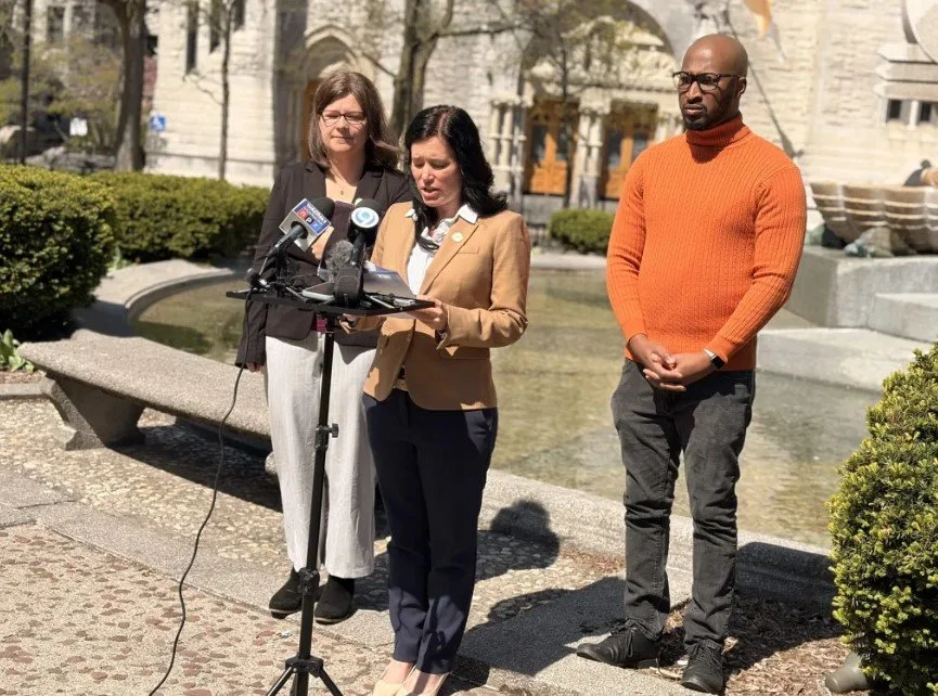 Three people standing outdoors in front of a fountain, with the woman in the beige blazer reading from a document at a microphone, and two others listening.