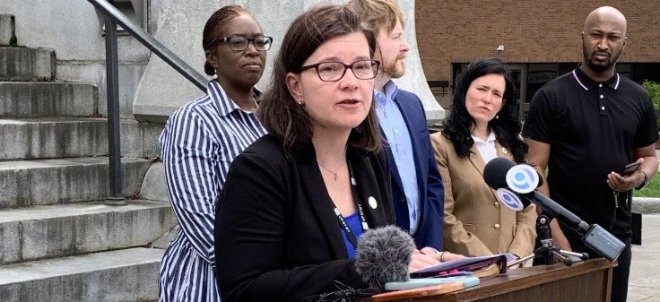 A woman speaking at a press conference with four people standing behind her, outdoors near a staircase.