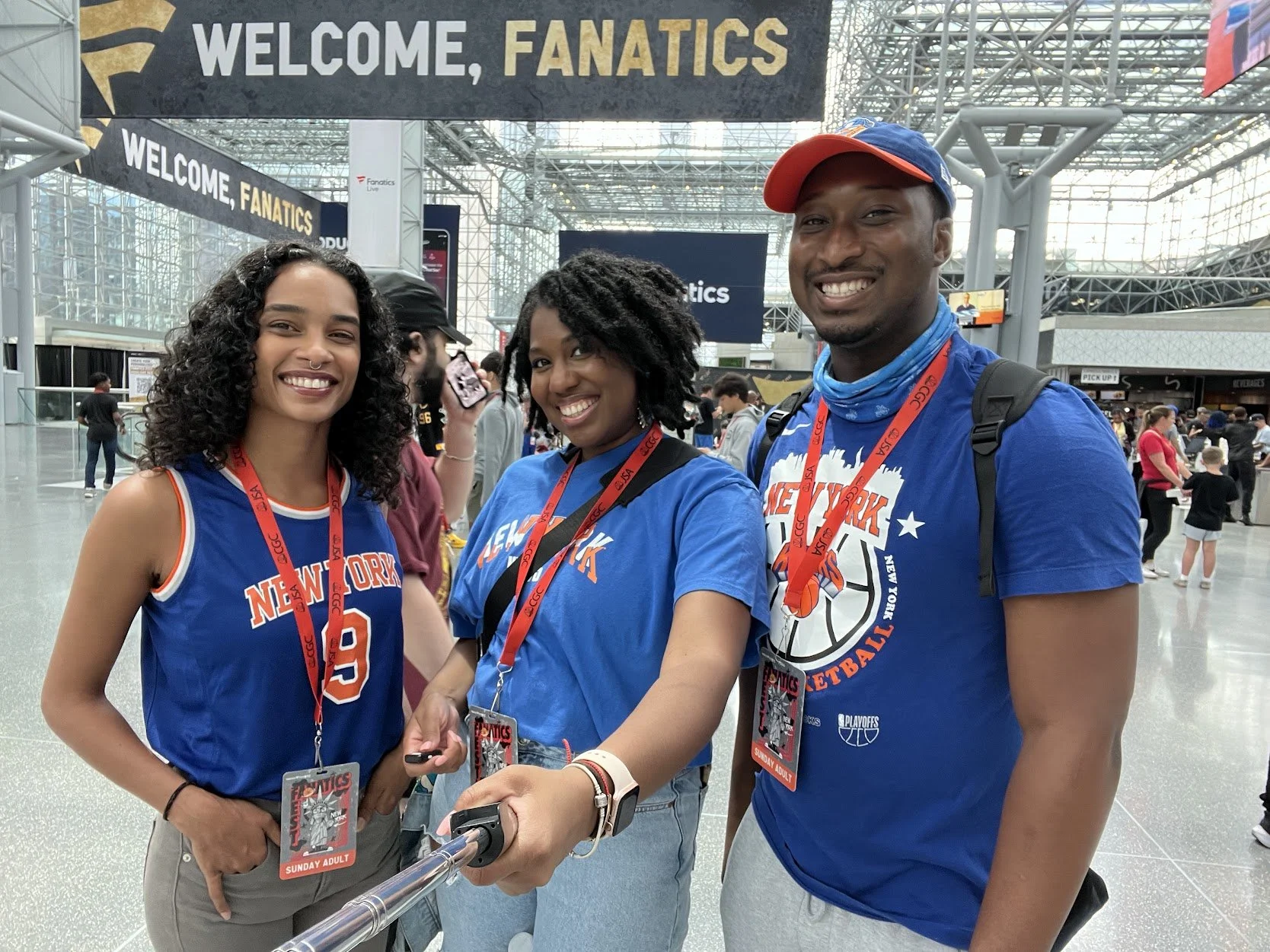 Three diverse people wearing New York Knicks basketball shirts and badges, taking a selfie together at a sports event, with a large