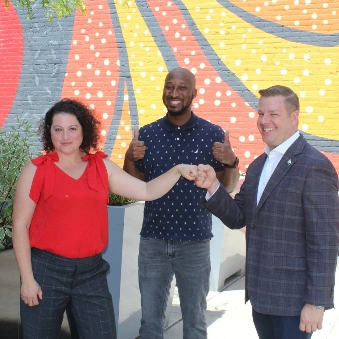 Three people, two men and one woman, are smiling and engaging in a fist bump outside in front of a colorful mural. The woman on the left has curly dark hair, wearing a red sleeveless top with bow details on the shoulders and dark checkered pants. The man in the middle is bald, has a beard, and is wearing a navy blue shirt with white polka dots and jeans. The man on the right has short blond hair, wearing a gray plaid blazer, white shirt, and dark pants.